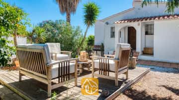 Muebles de patio de madera con cojines blancos sobre una terraza de madera frente a Villa Sofia, que presenta una fachada blanca, tejas de terracota y palmeras bajo un cielo azul despejado.