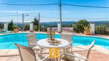 Zona de comedor al aire libre junto a la piscina en Villa Sofia, equipada con una mesa blanca de plástico, cuatro sillas a juego con cojines y un centro de mesa con linterna de mimbre, con vistas panorámicas a las colinas lejanas.