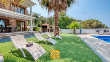 Soleado jardín en Villa Marcella que cuenta con tumbonas blancas sobre césped artificial, una palmera y una piscina de azulejos azules junto al exterior de ladrillo de la casa.