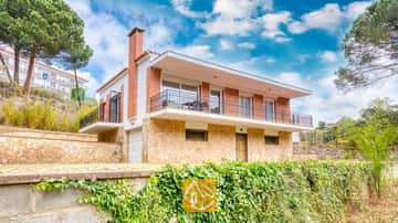 Vista exterior de Villa Lloret que muestra una casa de dos plantas con planta baja de piedra, piso superior de ladrillo y un balcón corrido bajo un cielo azul nublado.