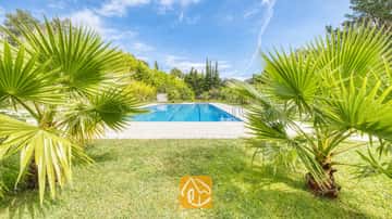 Piscina exterior comunitaria en Villa Lloret enmarcada por palmeras verdes, césped y árboles bajo un cielo azul.