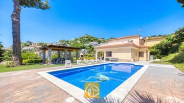 Zona de piscina al aire libre en Villa Paris con aguas cristalinas, tumbonas en el patio adoquinado y una pérgola cubierta junto a la casa de dos plantas de color beis.