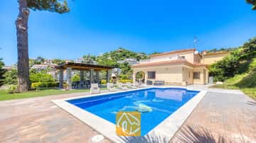 Zona de piscina al aire libre en Villa Paris con aguas cristalinas, tumbonas en el patio adoquinado y una pérgola cubierta junto a la casa de dos plantas de color beis.