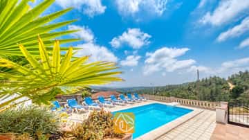 Piscina privada con tumbonas azules y una hoja de palmera en primer plano en Villa Santa Maria, bajo un cielo azul con nubes dispersas.