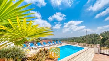 Piscina privada con tumbonas azules y una hoja de palmera en primer plano en Villa Santa Maria, bajo un cielo azul con nubes dispersas.