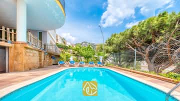 Piscina exterior en Villa Valentina con aguas cristalinas, rodeada de pavimento de baldosas, cinco tumbonas y pinos bajo un cielo azul.