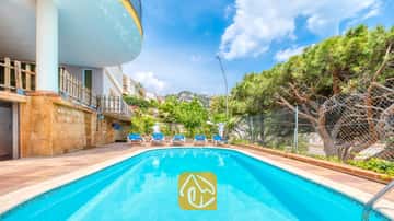 Piscina exterior en Villa Valentina con aguas cristalinas, rodeada de pavimento de baldosas, cinco tumbonas y pinos bajo un cielo azul.