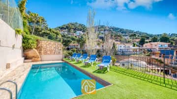 Piscina privada con tumbonas azules sobre una terraza de césped artificial verde en Villa Donna, con vistas a las casas en la ladera bajo un cielo azul despejado.