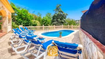 Piscina exterior privada en Villa Sarai rodeada de tumbonas azules, suelo de patio de piedra y árboles verdes bajo un cielo azul despejado