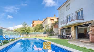 Piscina privada y terraza en Villa Nicky, con tumbonas azules sobre césped artificial junto a una villa blanca de dos plantas con pilares de piedra y puertas corredizas de cristal.