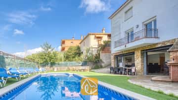 Piscina privada y terraza en Villa Nicky, con tumbonas azules sobre césped artificial junto a una villa blanca de dos plantas con pilares de piedra y puertas corredizas de cristal.