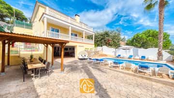 Piscina privada en Villa Ashley con aguas cristalinas y tumbonas blancas. La villa, de color amarillo, cuenta con balcones de balaustrada blanca y una pérgola de madera para disfrutar de comidas al aire libre sobre un suelo de piedra.