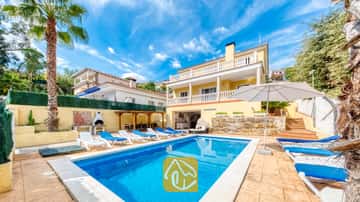 Piscina privada en Villa Ashley rodeada de tumbonas azules, una sombrilla blanca y un edificio amarillo de dos plantas con barandillas blancas en los balcones.