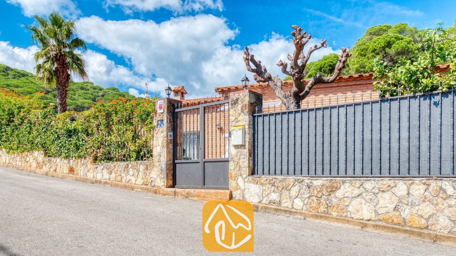 Fachada exterior y puerta de entrada metálica de Villa Alba, que presenta muros de piedra, una palmera alta y una calle adoquinada bajo un cielo azul.