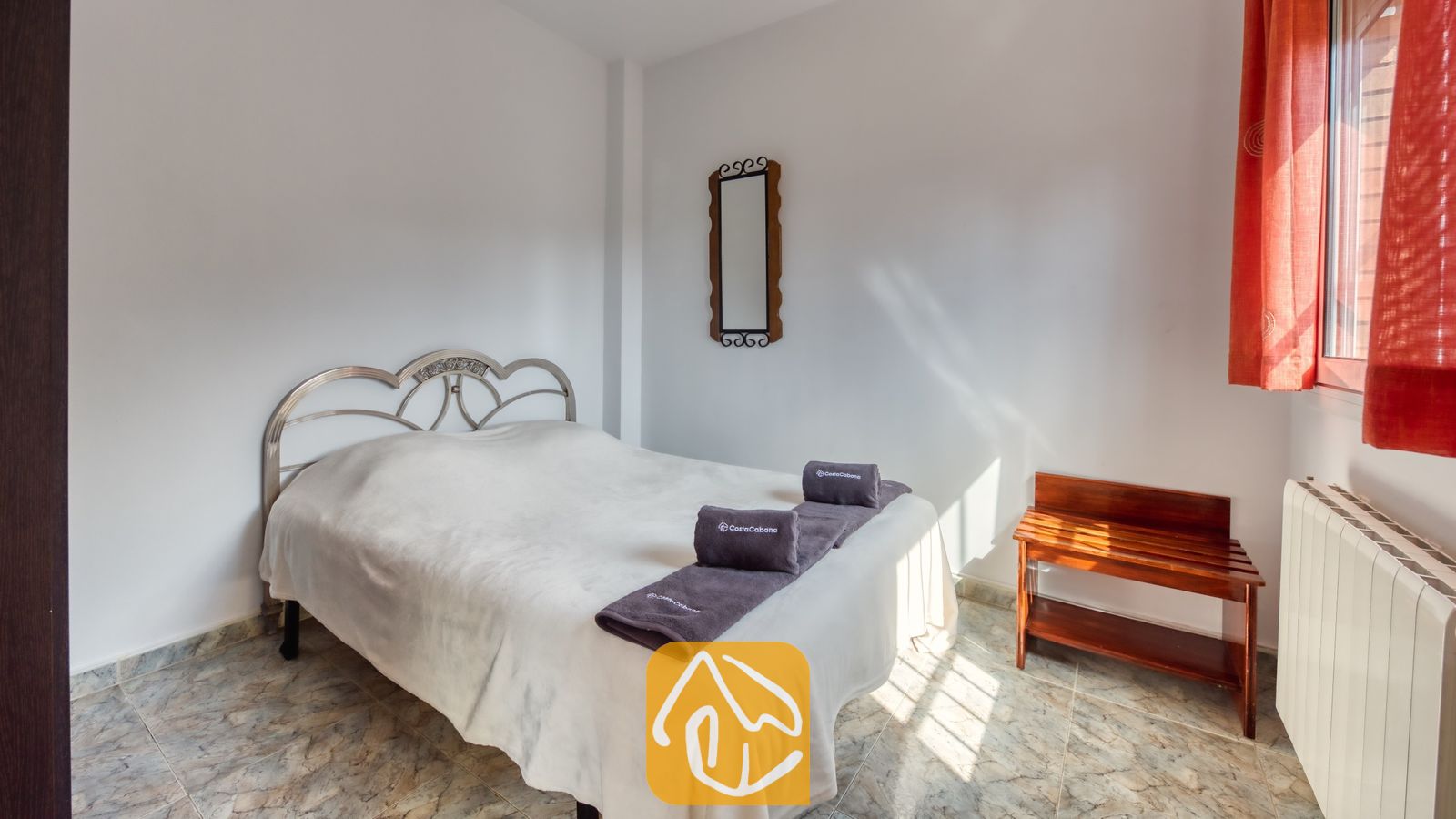 Dormitorio con una cama blanca, toallas grises dobladas, un cabecero de metal, cortinas rojas y un soporte para maletas de madera.