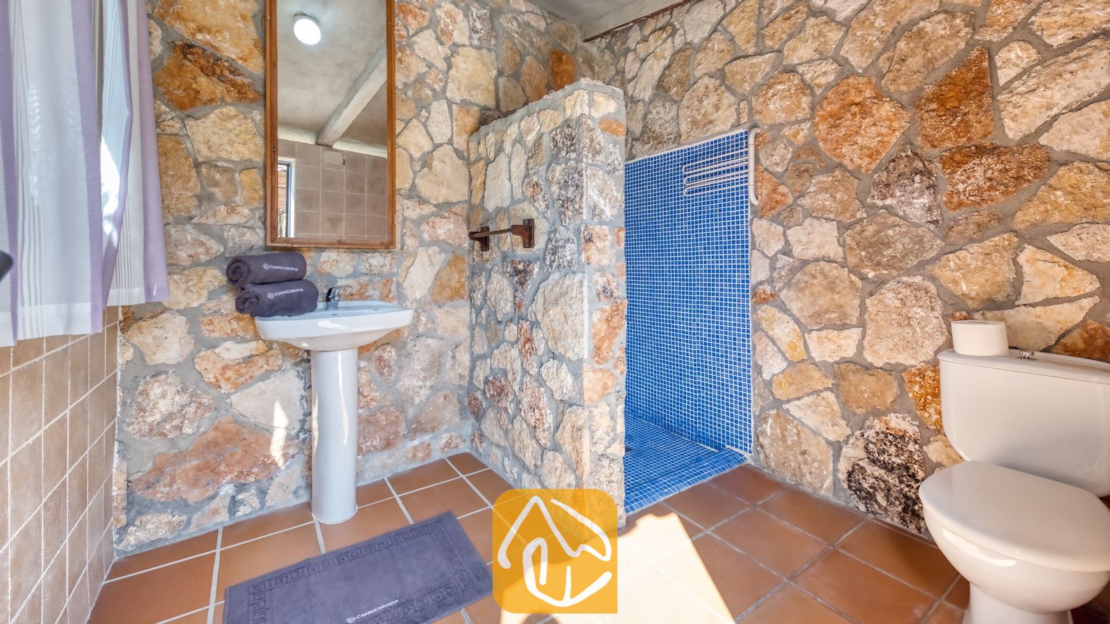 Cuarto de baño con paredes de piedra rústica, lavabo clásico con columna blanca, ducha acristalada con azulejos de mosaico en tonos azules y suelo de baldosas marrones.