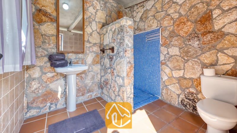 Cuarto de baño con paredes de piedra rústica, lavabo clásico con columna blanca, ducha acristalada con azulejos de mosaico en tonos azules y suelo de baldosas marrones.