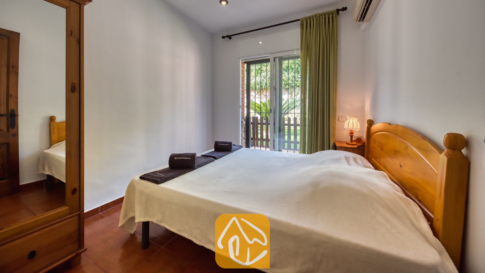 Dormitorio con cama doble de madera, armario a juego con espejo, toallas oscuras enrolladas y puerta corredera de cristal con acceso al balcón.
