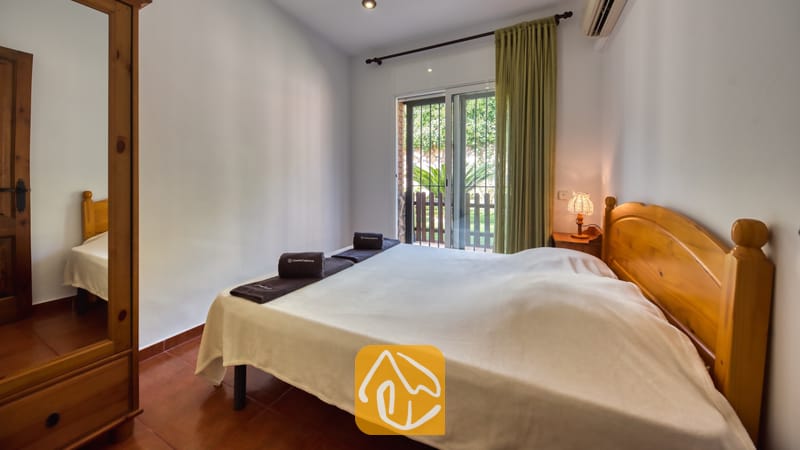 Dormitorio con cama doble de madera, armario a juego con espejo, toallas oscuras enrolladas y puerta corredera de cristal con acceso al balcón.