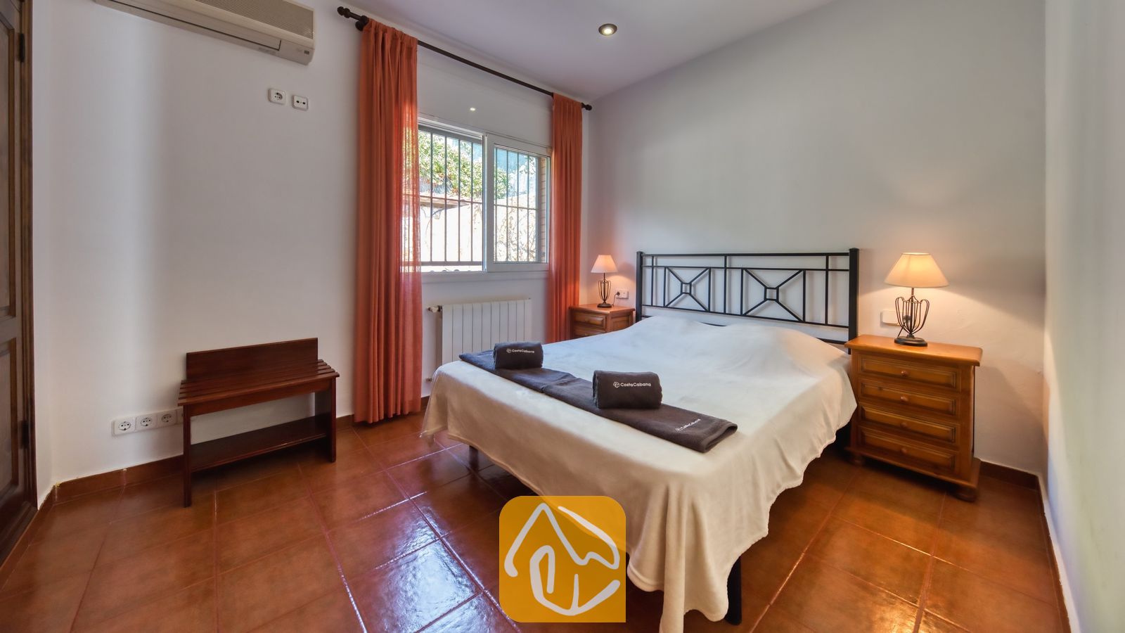 Dormitorio con cama doble, estructura de metal oscuro, cortinas naranjas que enmarcan la ventana y dos mesitas de noche de madera con lámparas.