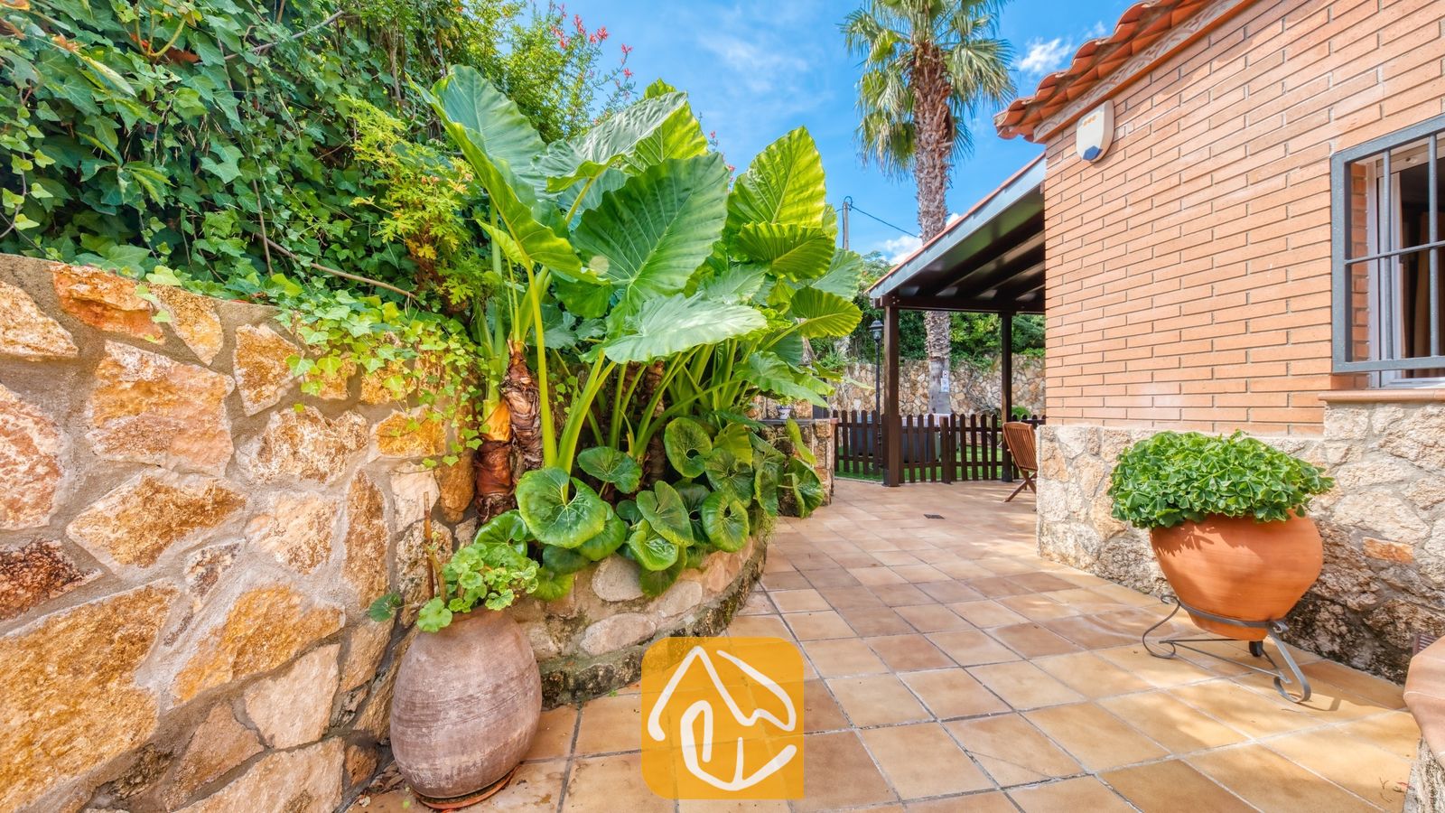 Patio exterior de Villa Alba que cuenta con un suelo de baldosas pavimentadas, grandes plantas tropicales verdes y un muro de piedra rústica.