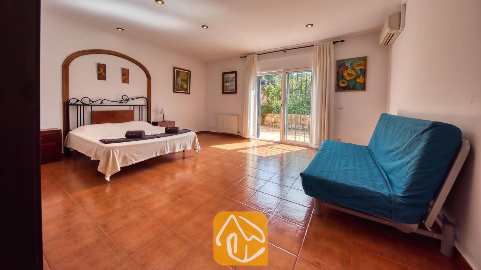Amplio dormitorio en Villa Alba con cama doble, estructura de metal oscuro, sofá azul y puertas correderas de cristal con acceso a una terraza exterior.