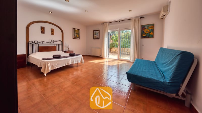 Amplio dormitorio en Villa Alba con cama doble, estructura de metal oscuro, sofá azul y puertas correderas de cristal con acceso a una terraza exterior.