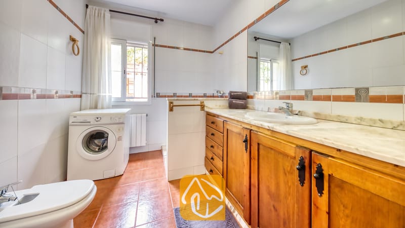 Baño luminoso de azulejos que cuenta con un mueble de madera con encimera de mármol, una lavadora blanca y suelo de baldosas de terracota.