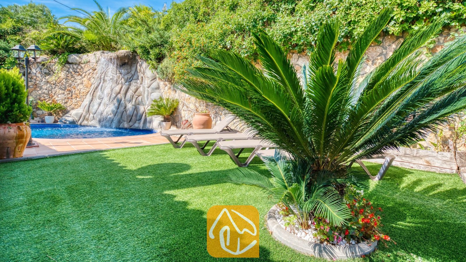 Frondoso y verde jardín en Villa Alba que cuenta con césped artificial, una palmera cícada destacada, tumbonas y una fuente de piedra con cascada.
