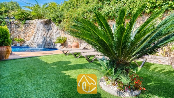Frondoso y verde jardín en Villa Alba que cuenta con césped artificial, una palmera cícada destacada, tumbonas y una fuente de piedra con cascada.