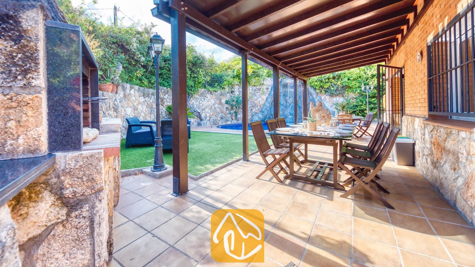 Terraza exterior cubierta para comer al aire libre en Villa Alba, equipada con una mesa y sillas de madera, suelo de baldosa y una zona de barbacoa de obra adyacente, con vistas al jardín y a la piscina