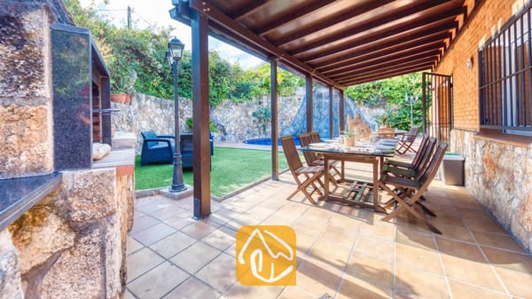 Terraza exterior cubierta para comer al aire libre en Villa Alba, equipada con una mesa y sillas de madera, suelo de baldosa y una zona de barbacoa de obra adyacente, con vistas al jardín y a la piscina