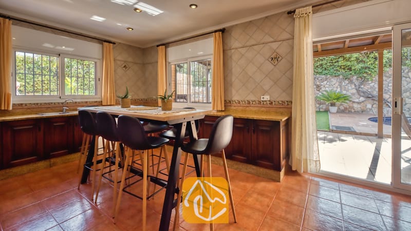 Zona de comedor de la cocina con una mesa alta de madera y taburetes de bar negros, suelo de baldosas de terracota, paredes alicatadas y una puerta de cristal abierta que da a una terraza exterior en Villa Alba.