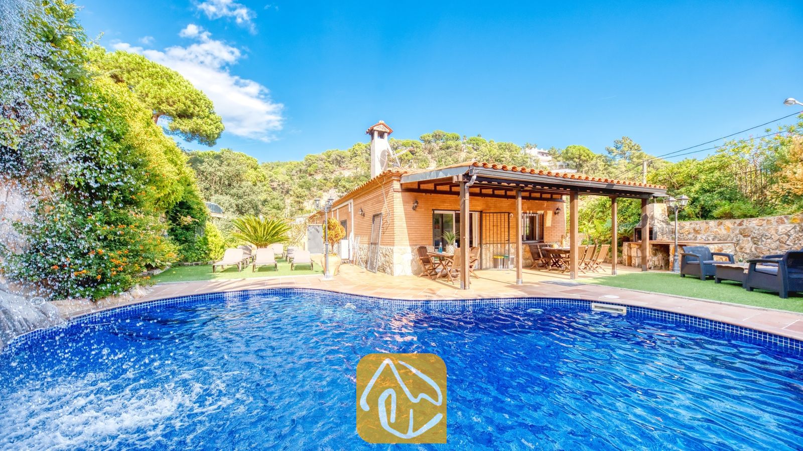 Piscina privada con cascada de piedra en Villa Alba, que cuenta con una casa de fachada de ladrillo, porche cubierto y tumbonas sobre césped artificial bajo un cielo azul.