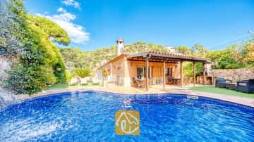 Piscina privada con cascada de piedra en Villa Alba, que cuenta con una casa de fachada de ladrillo, porche cubierto y tumbonas sobre césped artificial bajo un cielo azul.