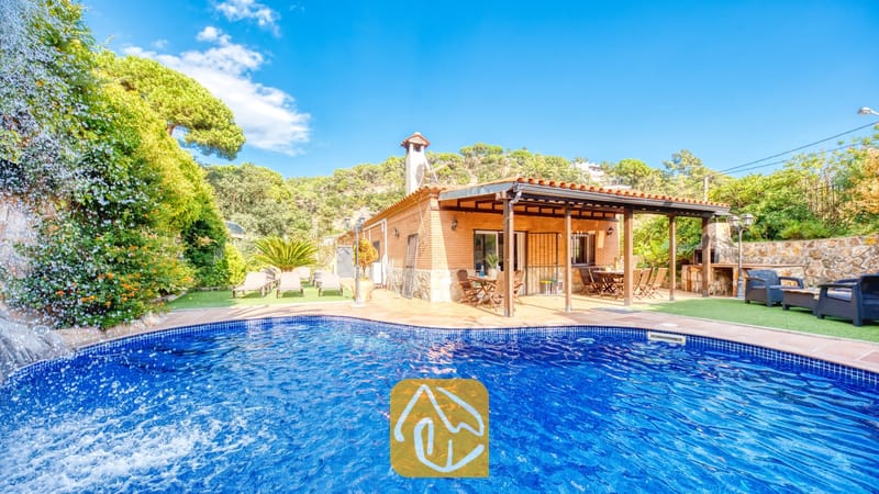 Piscina privada con cascada de piedra en Villa Alba, que cuenta con una casa de fachada de ladrillo, porche cubierto y tumbonas sobre césped artificial bajo un cielo azul.