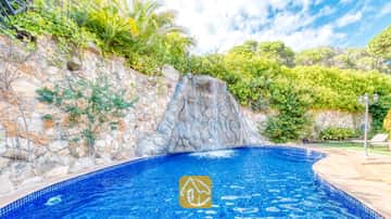 Cascada de roca que fluye hacia una piscina de azulejos azules en Villa Alba, rodeada de muros de contención de piedra y frondoso follaje bajo un cielo parcialmente nublado.