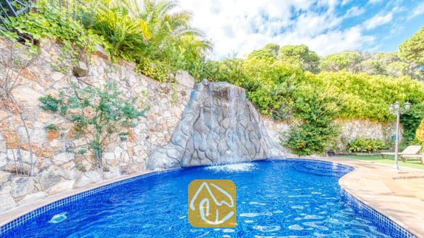 Cascada de roca que fluye hacia una piscina de azulejos azules en Villa Alba, rodeada de muros de contención de piedra y frondoso follaje bajo un cielo parcialmente nublado.