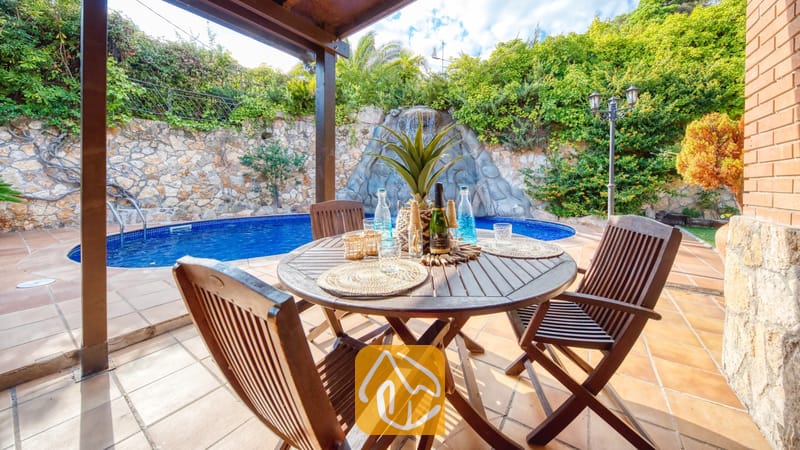 Mesa de comedor de madera y sillas en un patio de losetas en Villa Alba, con una piscina azul y una cascada de roca de fondo.