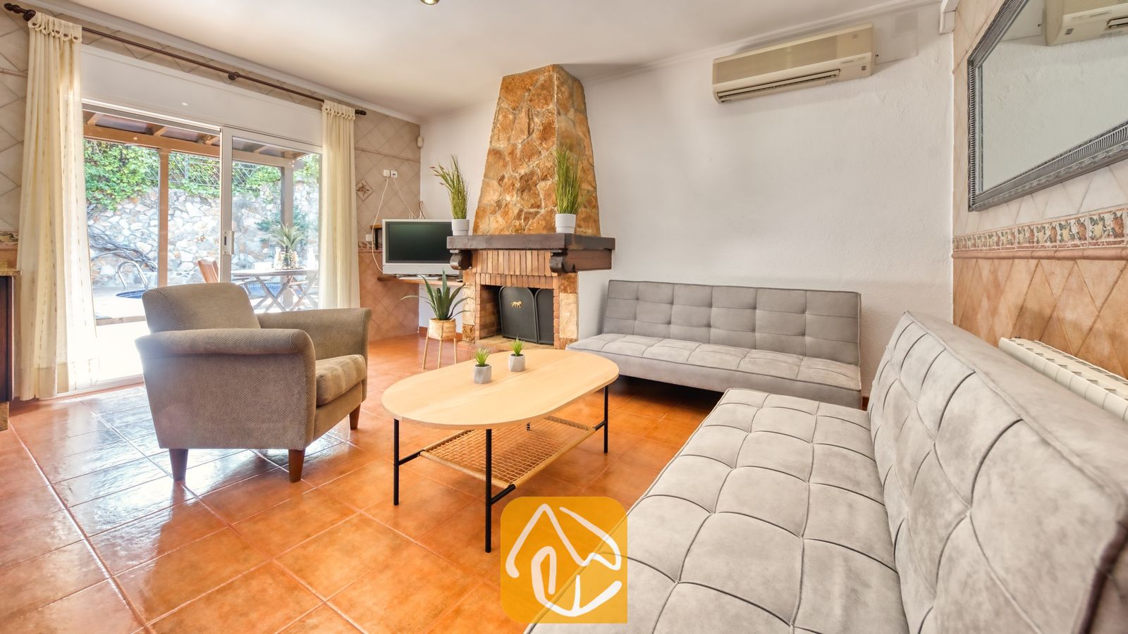 Salón de Villa Alba con dos sofás capitoné grises, un sillón, una mesa de centro ovalada de madera y una chimenea de piedra.