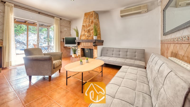 Salón de Villa Alba con dos sofás capitoné grises, un sillón, una mesa de centro ovalada de madera y una chimenea de piedra.