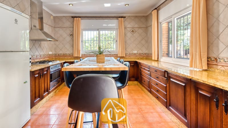 Amplia cocina equipada con gabinetes de madera oscura, encimeras de granito, una mesa de comedor central de madera con sillas negras y suelos de terracota.