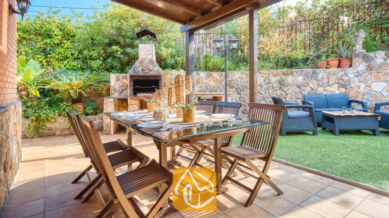 Patio comedor exterior cubierto en Villa Alba, equipado con una mesa de madera con tapa de cristal, sillas plegables y una barbacoa de piedra integrada al fondo.