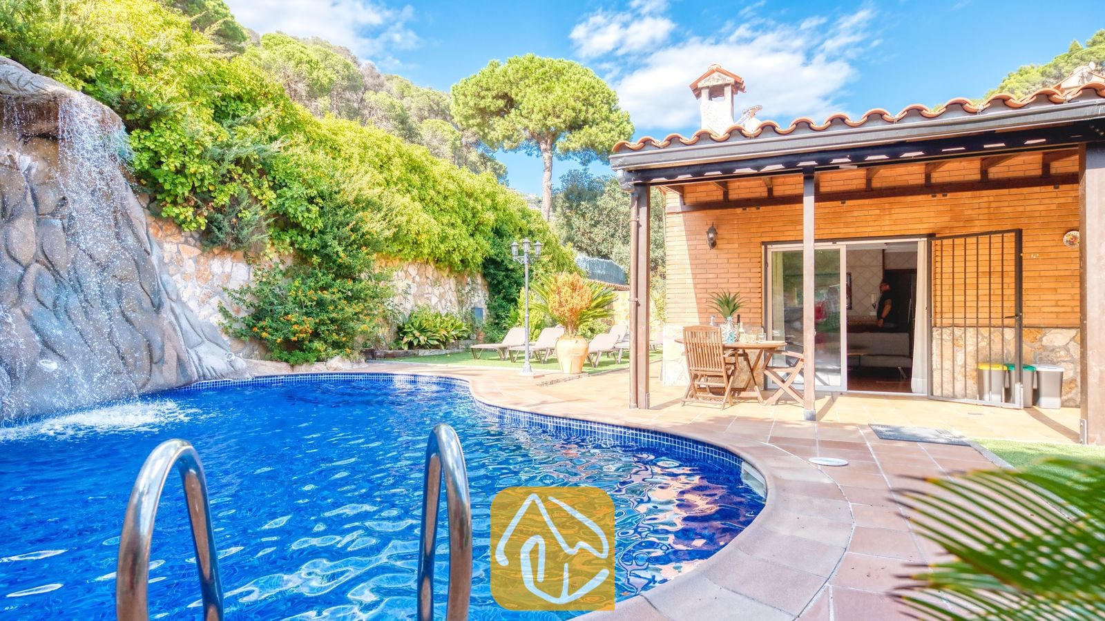 Piscina privada con una cascada de roca de piedra en Villa Alba, que muestra un patio exterior de ladrillo con muebles de comedor de madera y tejado de tejas.
