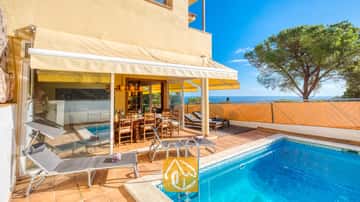 Piscina y terraza en Villa Santa Cristina con suelos de terracota, tumbonas grises y un comedor exterior de madera bajo un toldo amarillo, con vistas al mar.