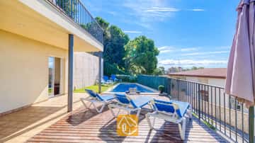 Soleada terraza de madera en Villa Mauri con tumbonas azules, una piscina en la planta baja y una zona de patio cubierto bajo un cielo azul despejado