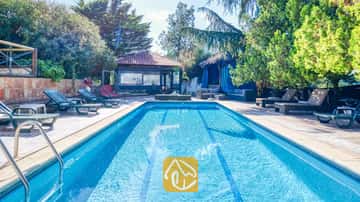 Piscina rectangular en Villa Can Bernardi rodeada de tumbonas verdes, árboles y un cenador exterior con tejado de tejas.