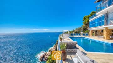 Piscina infinitaria al borde del acantilado en Villa Infinity, con tumbonas azules y amplias vistas al mar bajo un cielo despejado