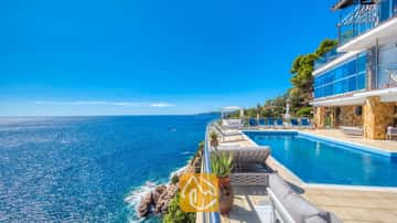 Piscina infinitaria al borde del acantilado en Villa Infinity, con tumbonas azules y amplias vistas al mar bajo un cielo despejado