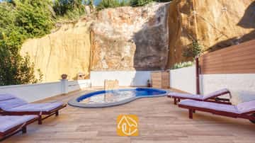 Piscina privada en Villa Blanca con azulejos de mosaico azul, ubicada en un patio adoquinado bordeado por una pared de roca alta y tumbonas de madera con cojines morados.